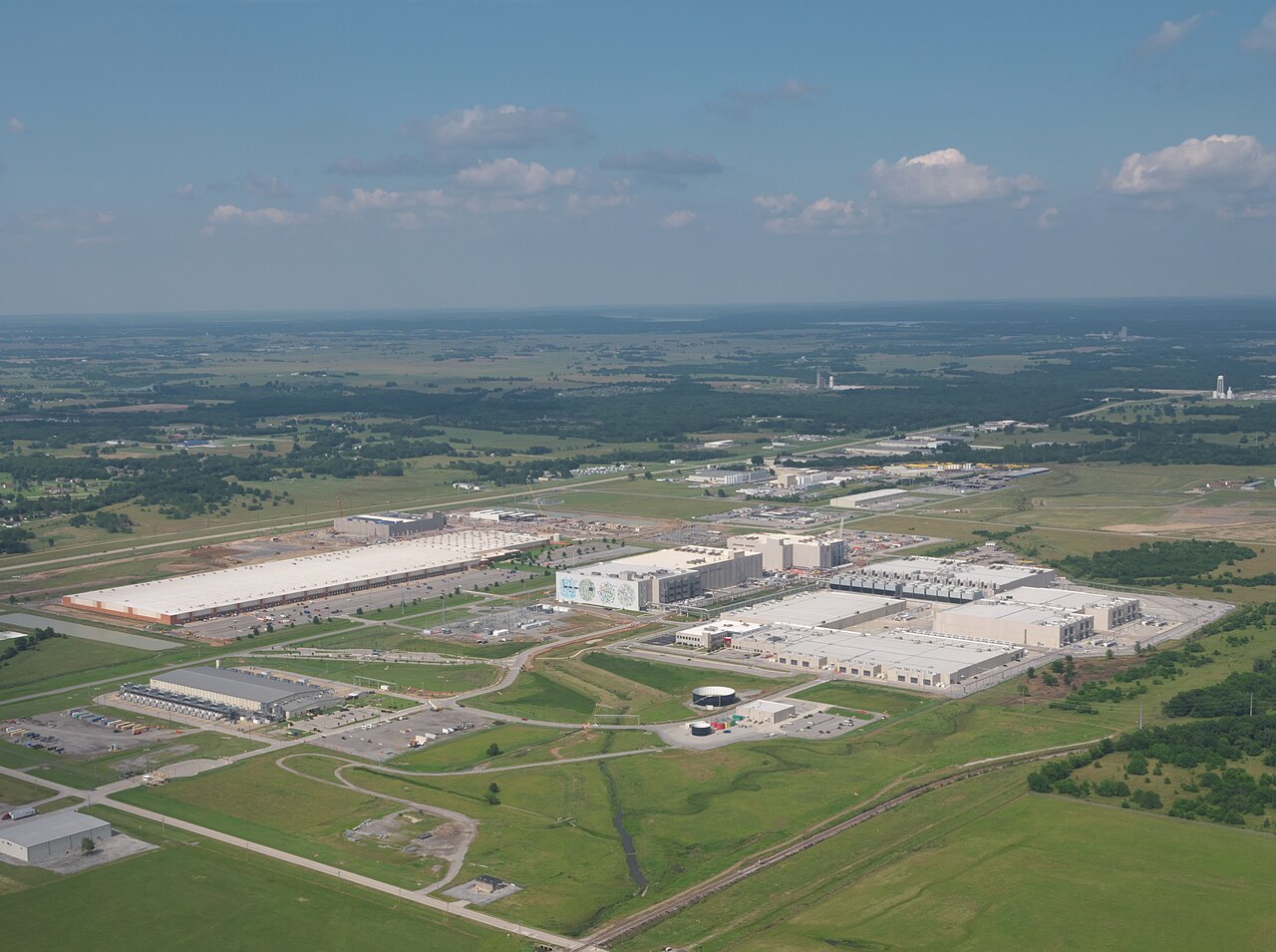 Aerial view of a data centre campus: several very long windowless halls with rooftop cooling plant, a circular water tank and haul roads, with green crop and pasture fields pressing right up to the fence on three sides and flat farmland running to the horizon.