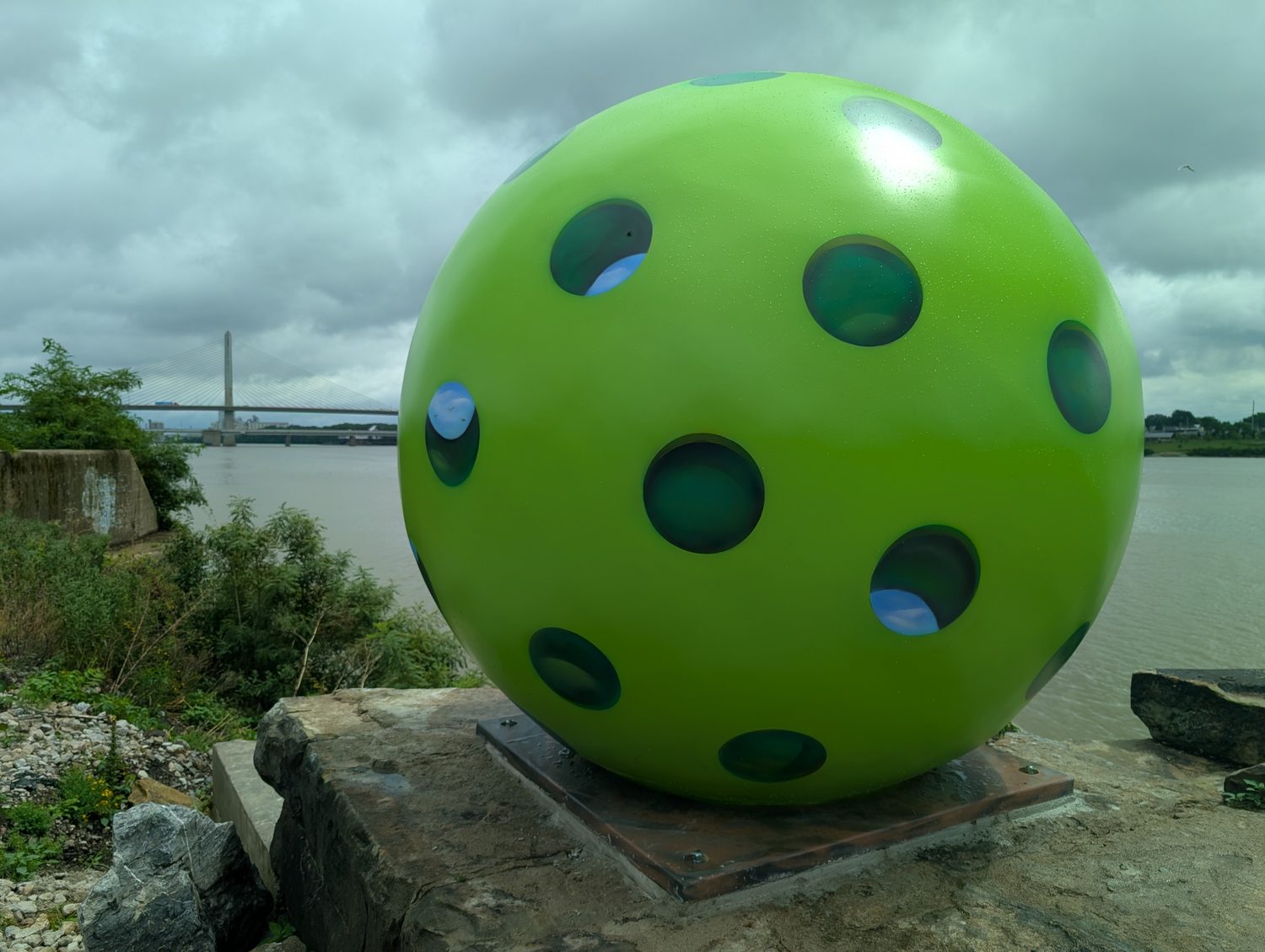 An eight-foot neon green pickleball bolted to a stone block on the riprap bank of the Maumee River, under a flat grey overcast, with the Veterans' Glass City Skyway standing in the distance at left. The ball's holes are painted rather than cut, and the ones catching the light show blue sky inside them.