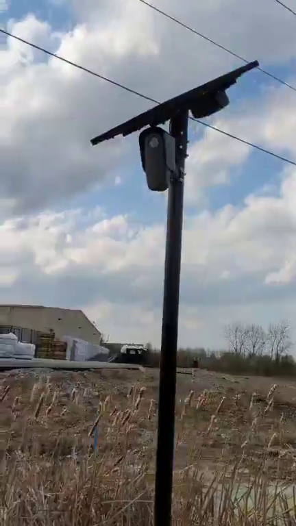A dark automatic licence plate reader camera and a solar panel mounted near the top of a slim post, seen against a bright cloudy sky with two power lines crossing behind it. Dry cattails in the foreground, a low warehouse and bare trees along the horizon.