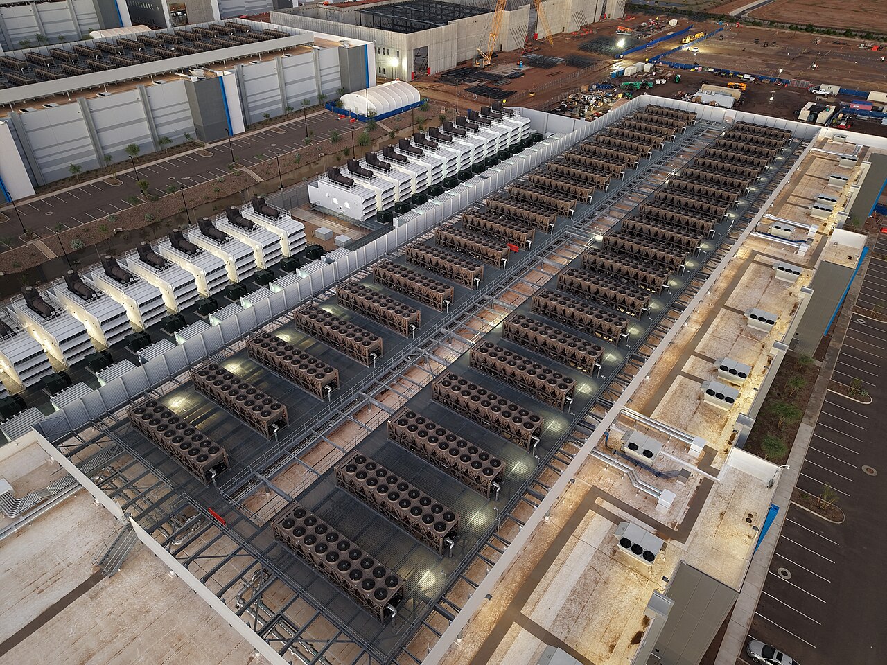 Night aerial view of a data centre roof: long rows of cooling units and banks of backup generators under floodlights, with a construction crane on an adjoining site. / Vista a&eacute;rea nocturna del techo de un centro de datos: largas filas de unidades de enfriamiento y bancos de generadores de respaldo bajo reflectores, con una gr&uacute;a de construcci&oacute;n en un terreno contiguo.