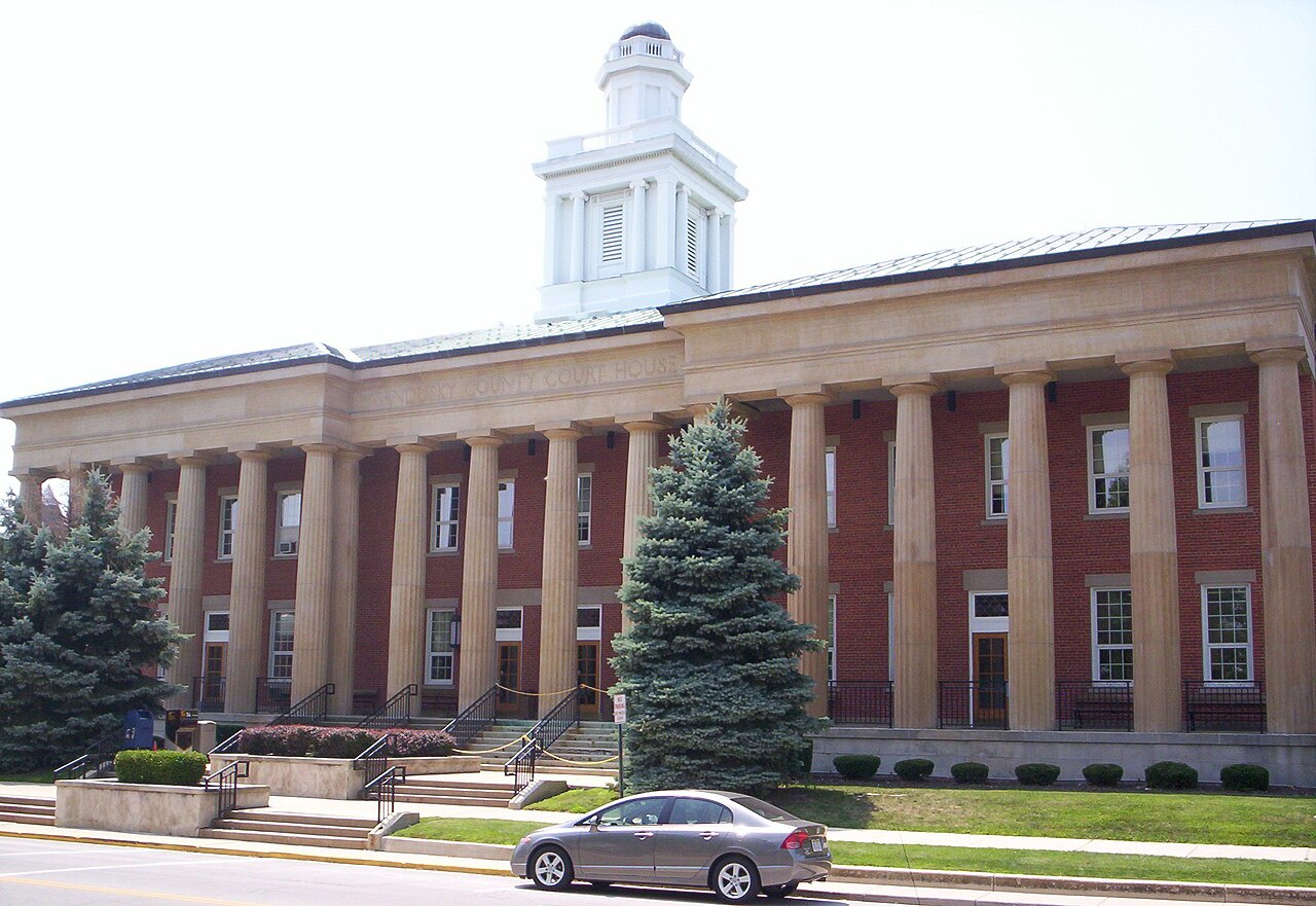 The Sandusky County Courthouse in Fremont, Ohio: a red brick building behind a long colonnade of pale stone columns, under a white cupola / El palacio de justicia del condado de Sandusky en Fremont, Ohio: un edificio de ladrillo rojo tras una larga columnata de piedra clara, bajo una cúpula blanca