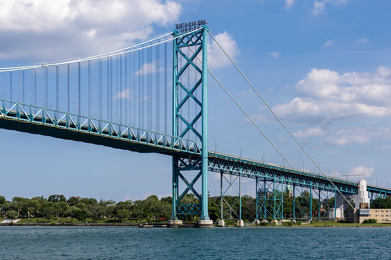The Ambassador Bridge seen from the Detroit River, its turquoise steel tower in the foreground and the deck running away toward the American shore. / El puente Ambassador visto desde el rio Detroit, con su torre de acero turquesa en primer plano.