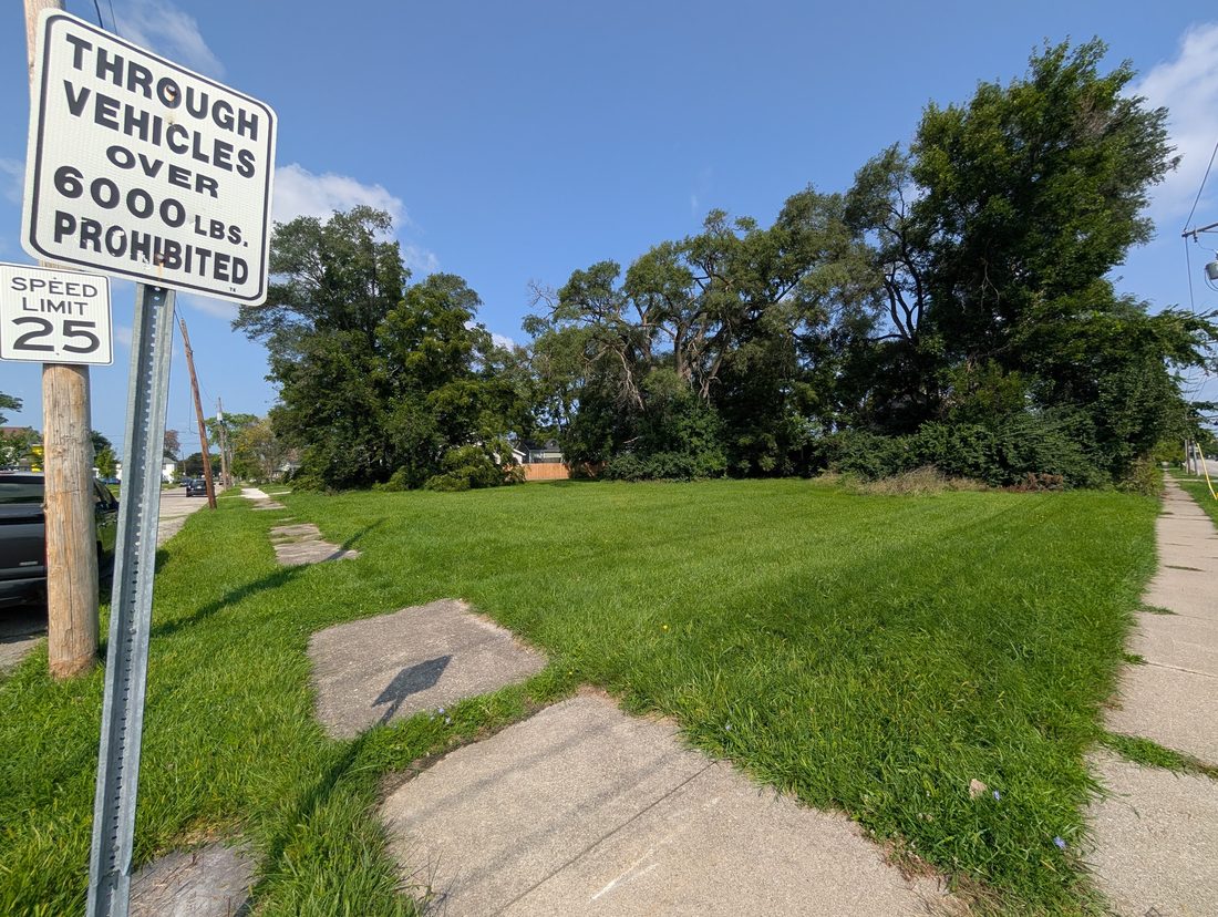 A wide mown vacant lot at a residential corner on Vance Street, with cracked sidewalk in the foreground and mature trees at the back of the lot.