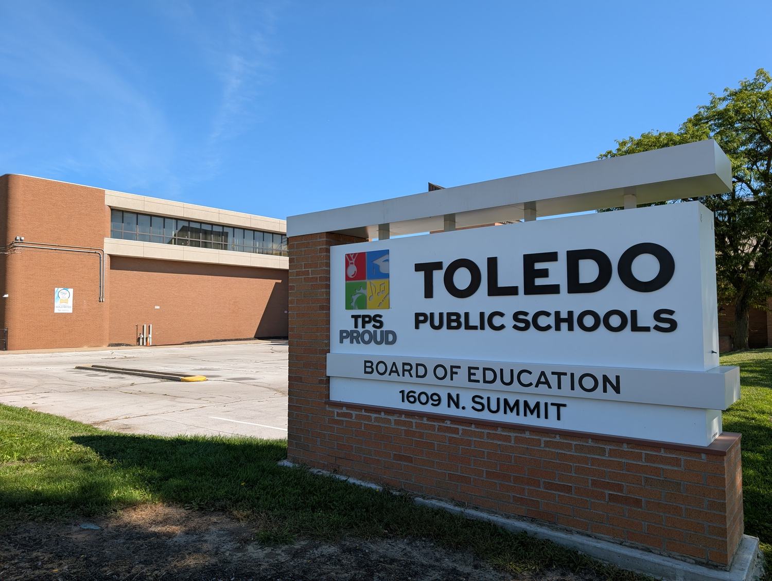 The Toledo Public Schools Board of Education sign at 1609 N. Summit St., reading TPS PROUD, with the district’s administration building behind it under a clear sky. / El letrero de la Junta de Educaci&oacute;n de las Escuelas P&uacute;blicas de Toledo en el 1609 de N. Summit St., con el edificio administrativo del distrito detr&aacute;s.