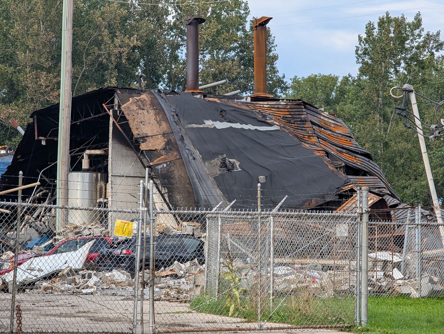 The collapsed rear of the Brent Industries plant on South Avenue: a charred, sagging roof membrane over buckled rust-orange steel, two stacks still standing, crushed cars and a spill of concrete block behind a chain-link fence / La parte trasera derrumbada de la planta de Brent Industries en South Avenue: una membrana de techo carbonizada y vencida sobre acero pandeado color óxido, dos chimeneas aún en pie, autos aplastados y bloques de concreto desparramados tras una cerca de alambre