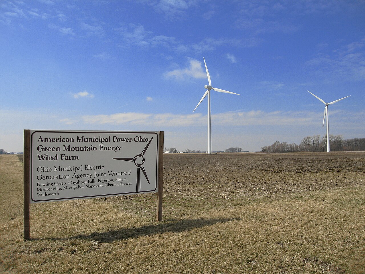 The entrance sign of the Bowling Green wind farm with two wind turbines standing behind it in a flat field. / El letrero de entrada del parque e&oacute;lico de Bowling Green con dos aerogeneradores detr&aacute;s, en un campo llano.