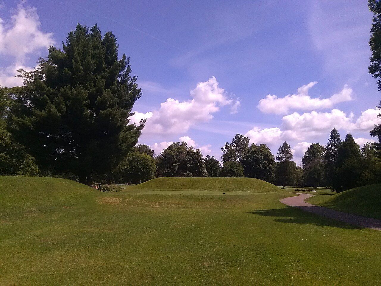 A grass mound inside the Octagon Earthworks in Newark, Ohio, in August 2016, with a mowed putting green at its foot and a paved golf-cart path to the right. / Un montículo de hierba dentro del Octágono de Newark, Ohio, en agosto de 2016, con un green de golf recortado a su pie y un sendero pavimentado para carritos a la derecha.