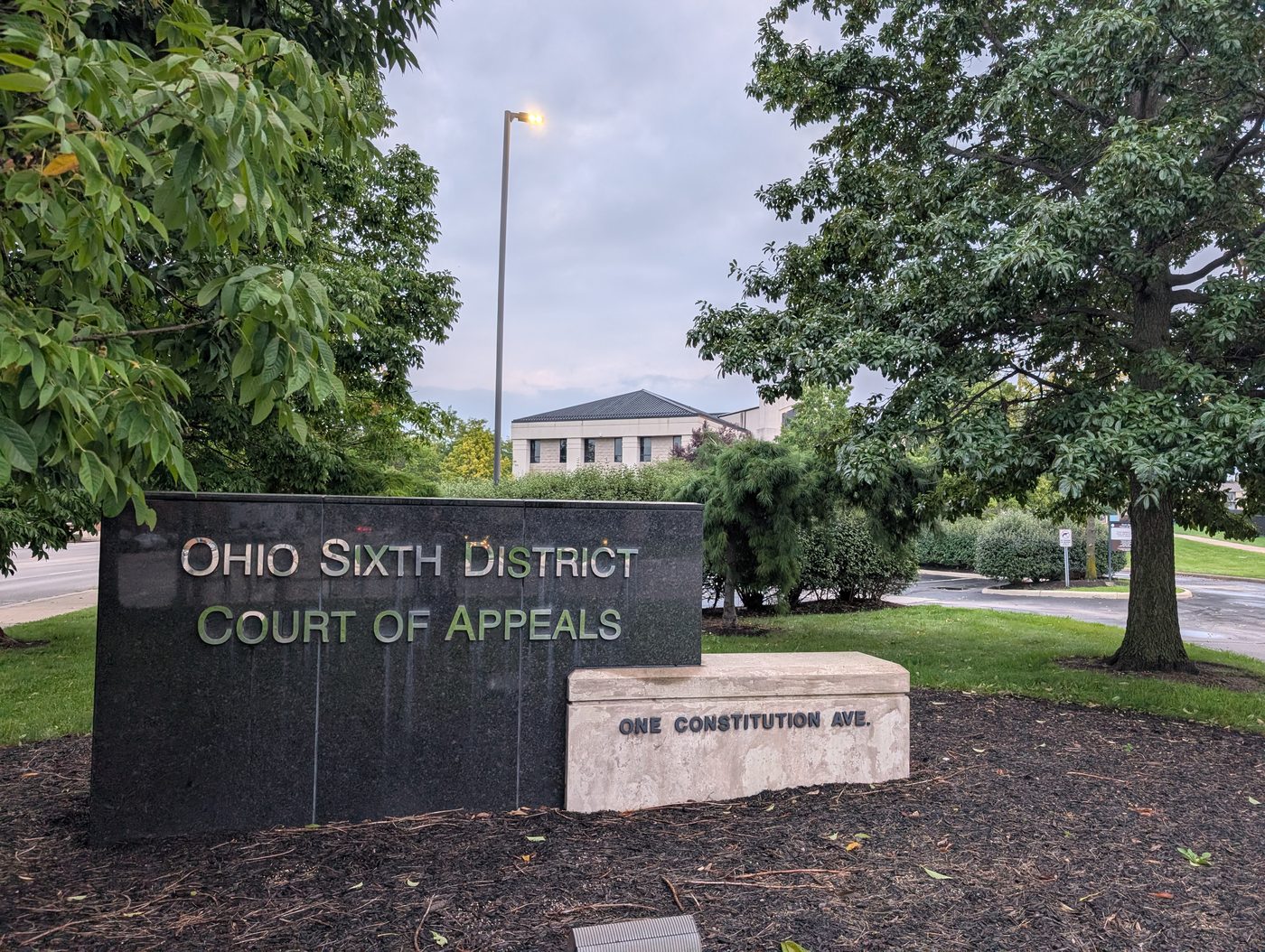 The monument sign outside the Ohio Sixth District Court of Appeals in Toledo, reading Ohio Sixth District Court of Appeals and One Constitution Ave., with the courthouse behind it through the trees. / El letrero monumental frente al Tribunal de Apelaciones del Sexto Distrito de Ohio, en Toledo, que dice Ohio Sixth District Court of Appeals y One Constitution Ave., con el edificio del tribunal detrás, entre los árboles.