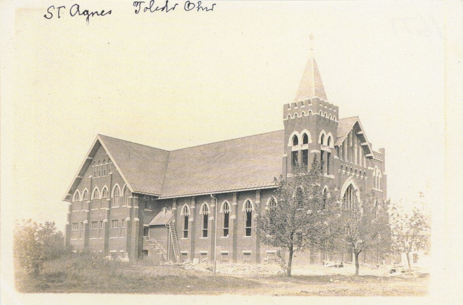 A sepia real-photo postcard of St. Agnes church in Toledo when it was new: brick Gothic Revival nave, corner tower with a crenellated top and short spire, a two-story school wing behind, construction debris and newly planted saplings in the yard, the words St. Agnes Toledo Ohio handwritten across the top. / Una postal fotográfica en sepia de la iglesia de St. Agnes en Toledo recién construida: nave neogótica de ladrillo, torre de esquina con remate almenado y aguja corta, un ala escolar de dos pisos detrás, escombros de obra y árboles recién plantados en el patio, y las palabras St. Agnes Toledo Ohio escritas a mano en la parte superior.