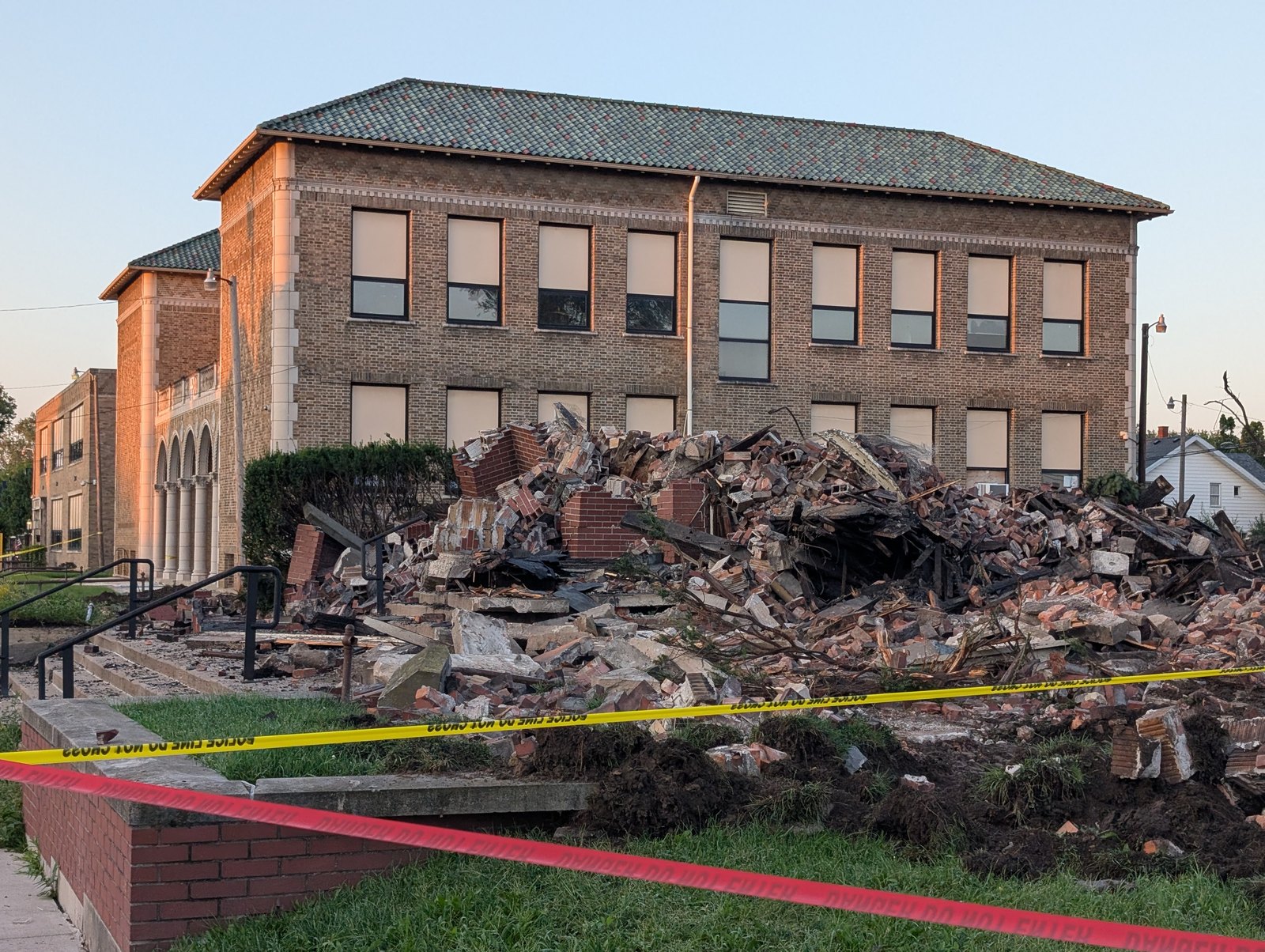 The rubble of the former St. Agnes church on Martha Avenue, a heap of brick and charred timber behind police tape, with the parish school building standing intact beside it / Los escombros de la antigua iglesia de St. Agnes en Martha Avenue, un montón de ladrillo y madera quemada tras la cinta policial, con el edificio de la escuela parroquial en pie a su lado