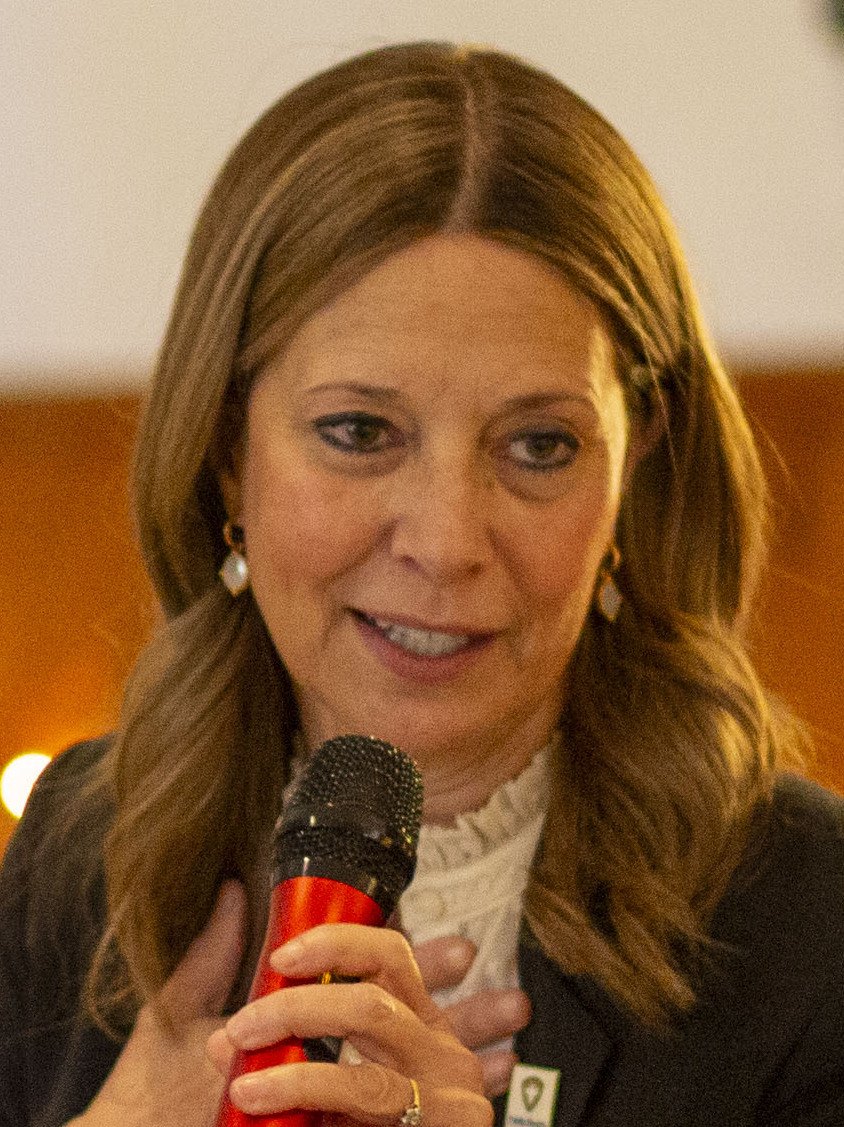 Amy Acton, in a dark jacket over a white blouse, speaking into a handheld microphone at an indoor event / Amy Acton, con chaqueta oscura sobre una blusa blanca, habla por un micrófono de mano en un evento bajo techo
