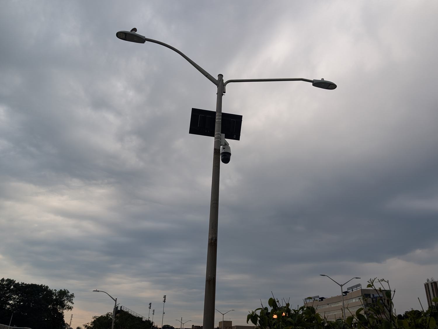 A Flock Safety pan-tilt-zoom dome camera and a small solar panel mounted on the arm of a streetlight, silhouetted against dark storm clouds. / Una camara domo Flock Safety con giro e inclinacion y un pequeno panel solar montados en el brazo de una farola, recortados contra nubes de tormenta oscuras.