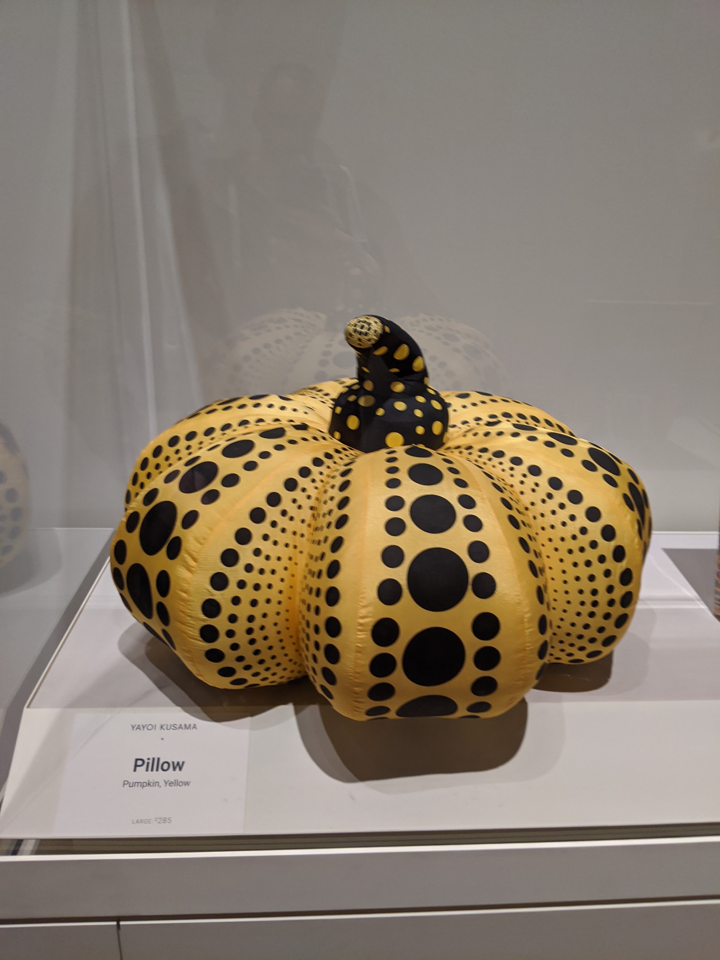 A yellow pumpkin covered in black dots, with a black dotted stem, in a museum vitrine above a card reading Yayoi Kusama, Pillow, Pumpkin Yellow, large, $285